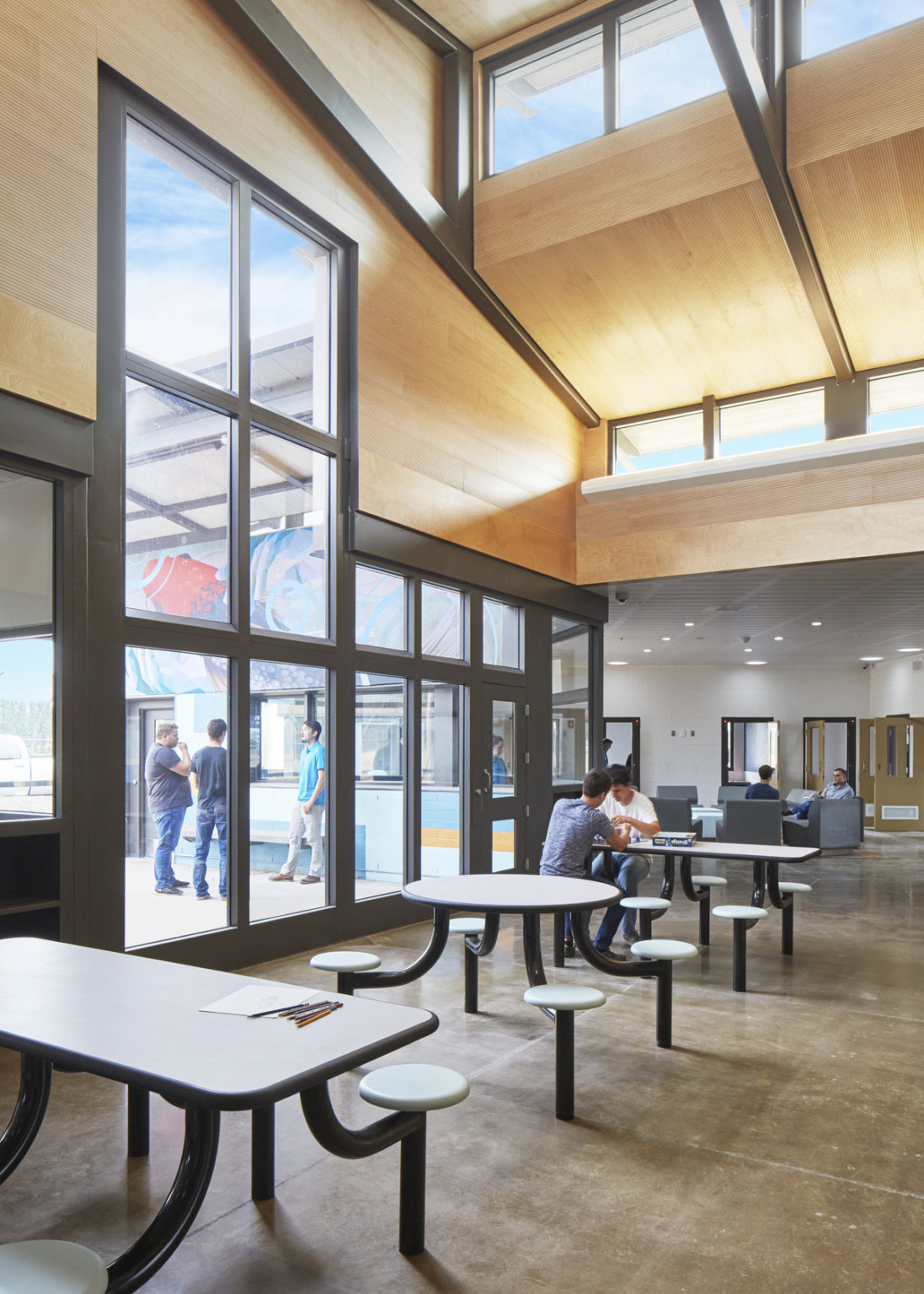 Wider view of double height wood hallway. Tables with attached chairs sit next to large windows. Beyond is a white hallway