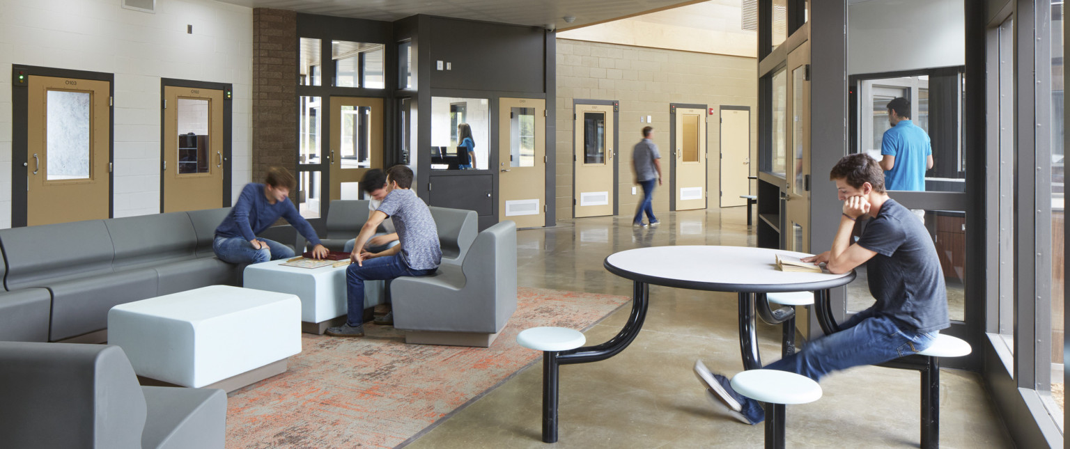 Partially carpeted seating area with tables in grey and white room at the end of white hallway. Light wooden doors line hall