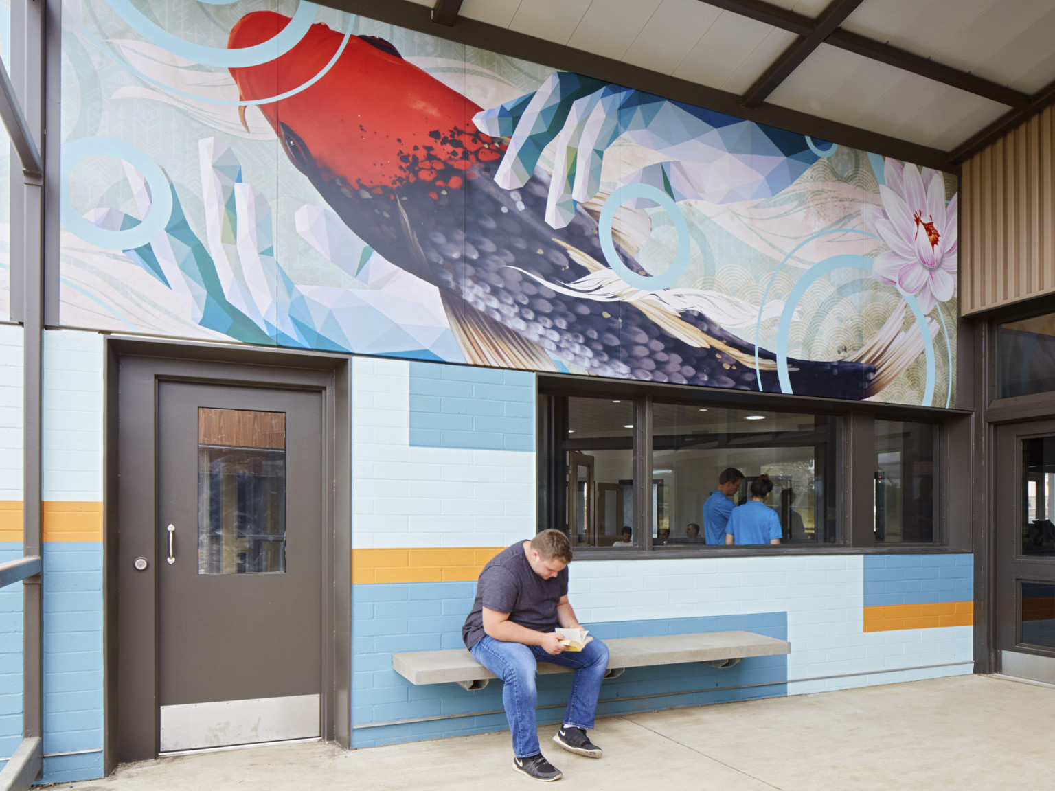 A grey bench against a blue and yellow colorblock wall with a window into an office. Above is a mural of a koi fish