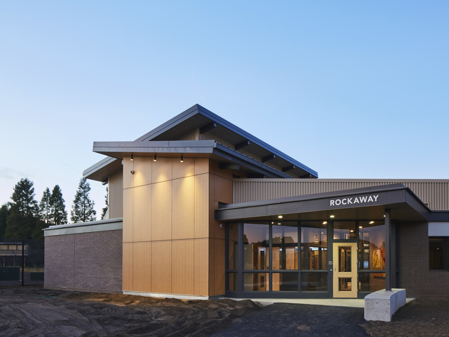 Exterior view of building entry with pointed overhang with wood panel base supported by black column anchored in cement bench