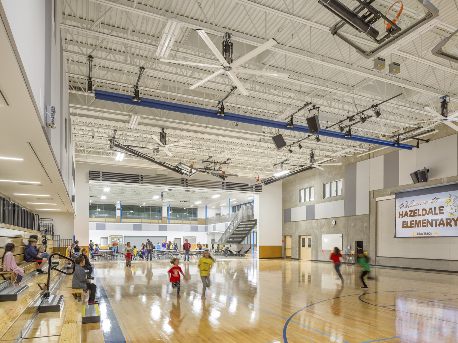 View of gymnasium in opposite direction. Flexible wall opened to cafeteria. Wood bleachers on left. Hazeldale Elementary sign