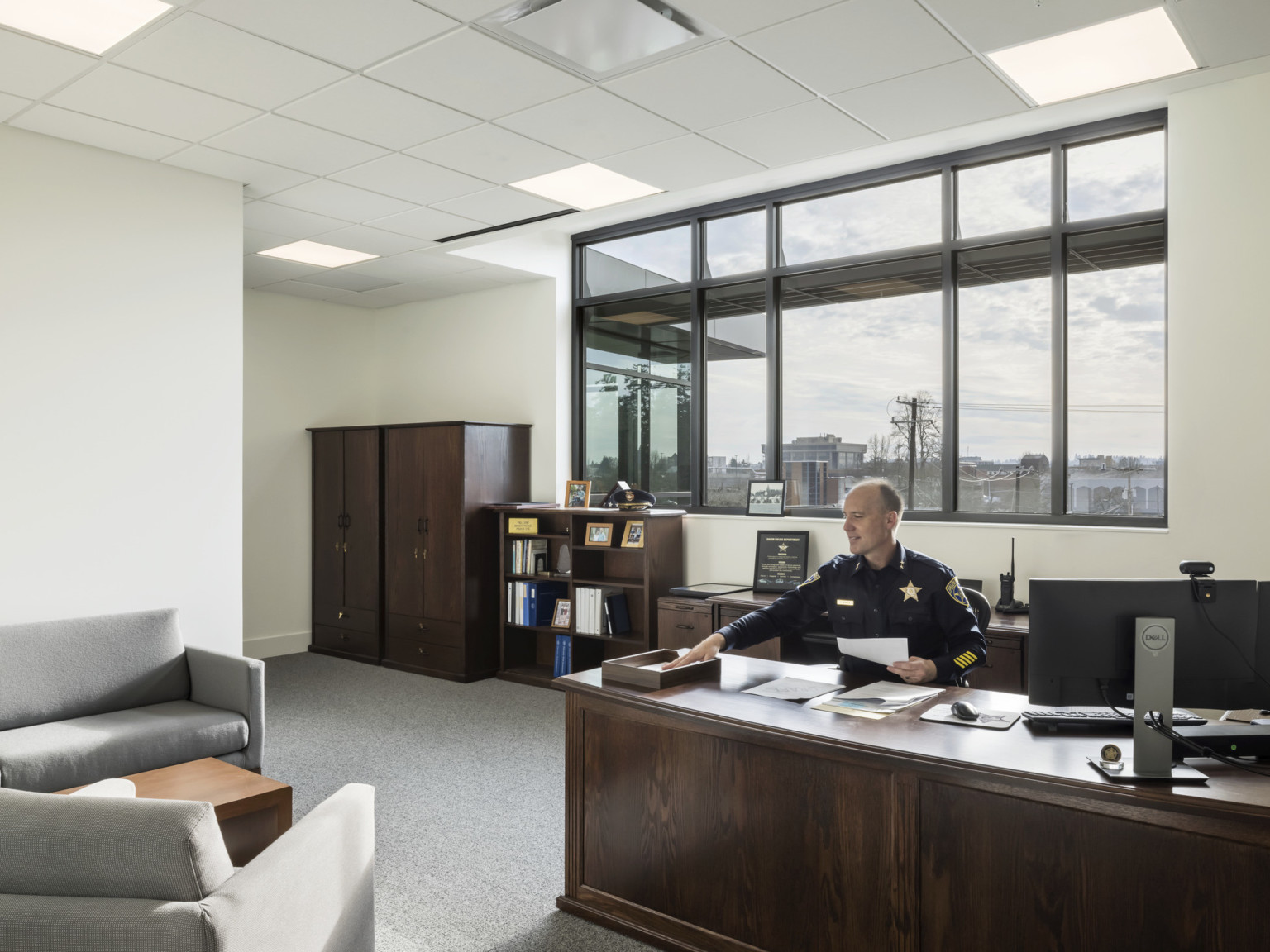 Office with man at dark wood desk with matching shelves in front of large window and a comfortable seating area in front