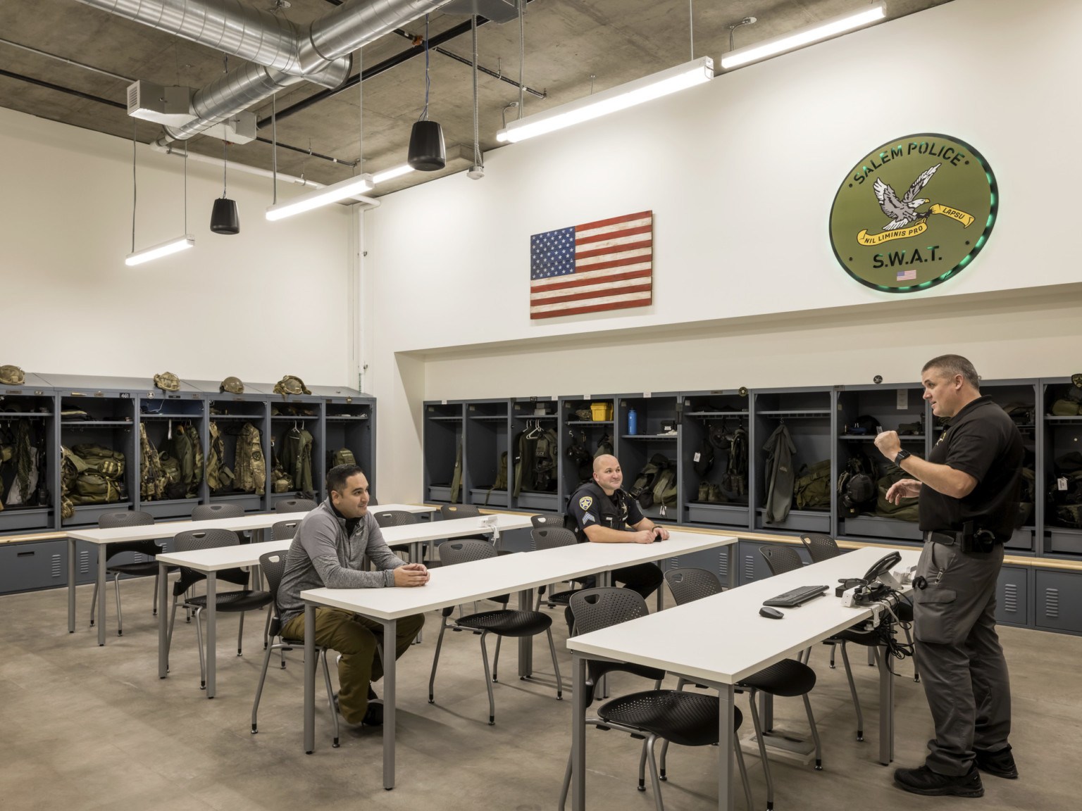 Training room with four tables with chairs with dark grey storage lining walls. Exposed ductwork and lighting on ceiling