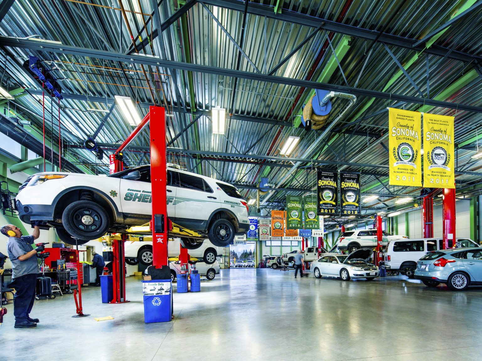 Interior with cars along side walls, some lifted by a red and yellow machine. Banners hang along center from paneled ceiling