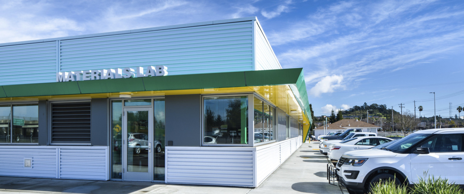Silver panel building with green and yellow awning with sign reading Materials Lab over glass door. Parking lot to the right