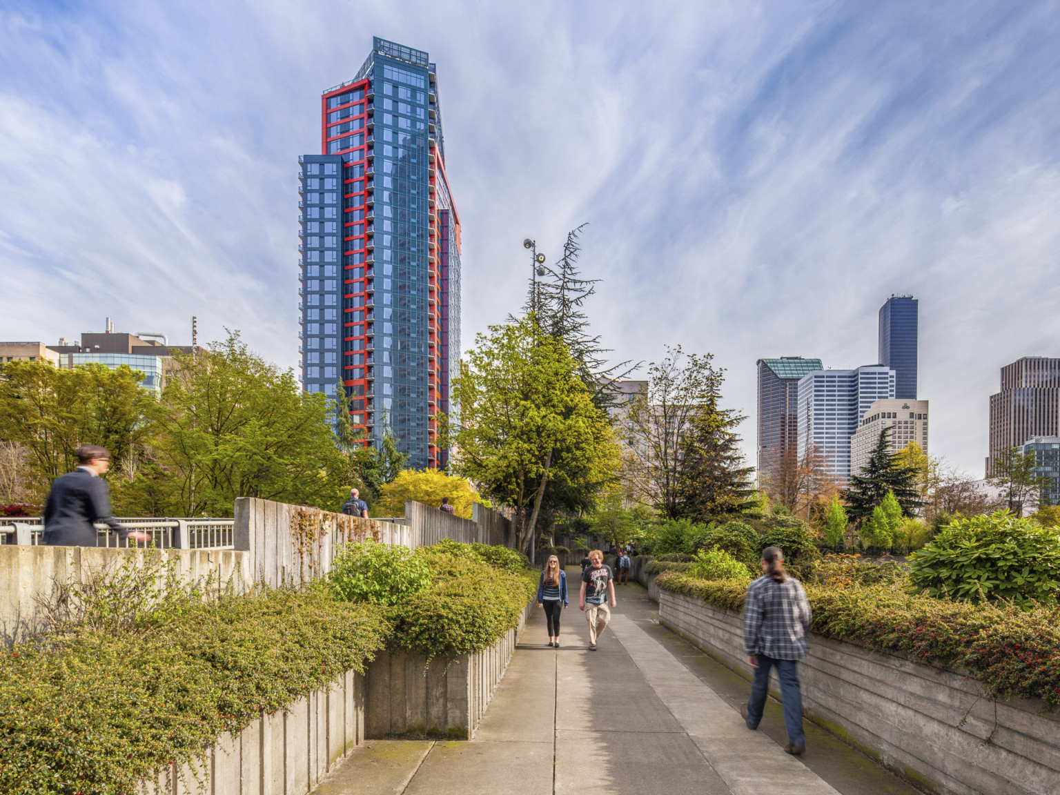 A layered glass building with red and blue wrapping facade, behind a tree lined walkway surrounded by skyscrapers