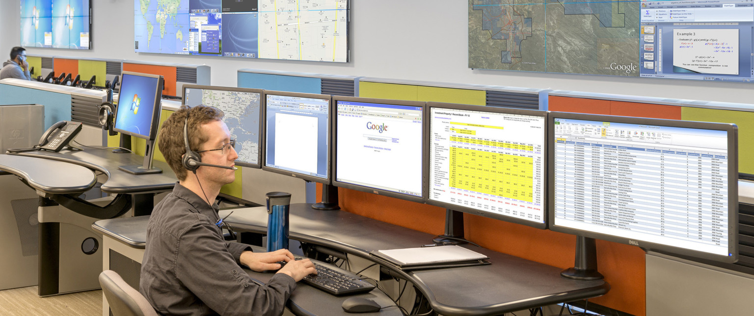 Man wearing headset sits at desk with 5 screens of spreadsheets and maps with colorful wall panels behind