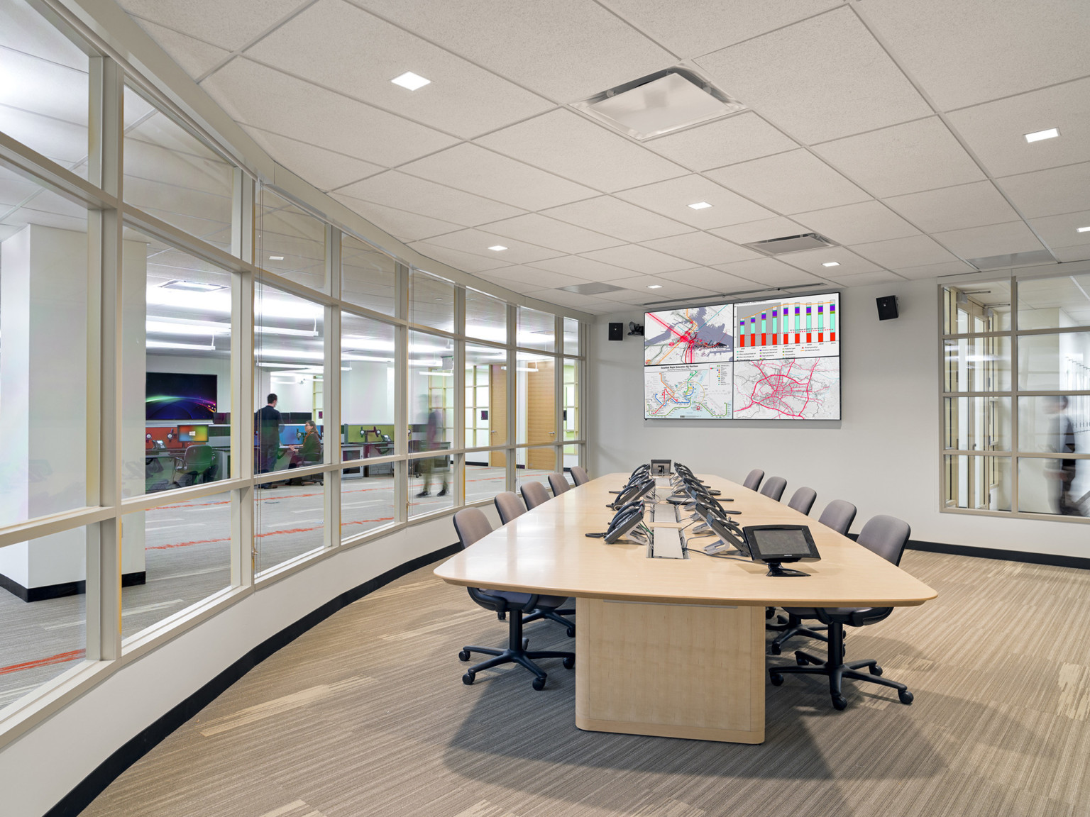 Conference room with long table with chairs and phones. Screens on opposite wall. Windows to hallway, left