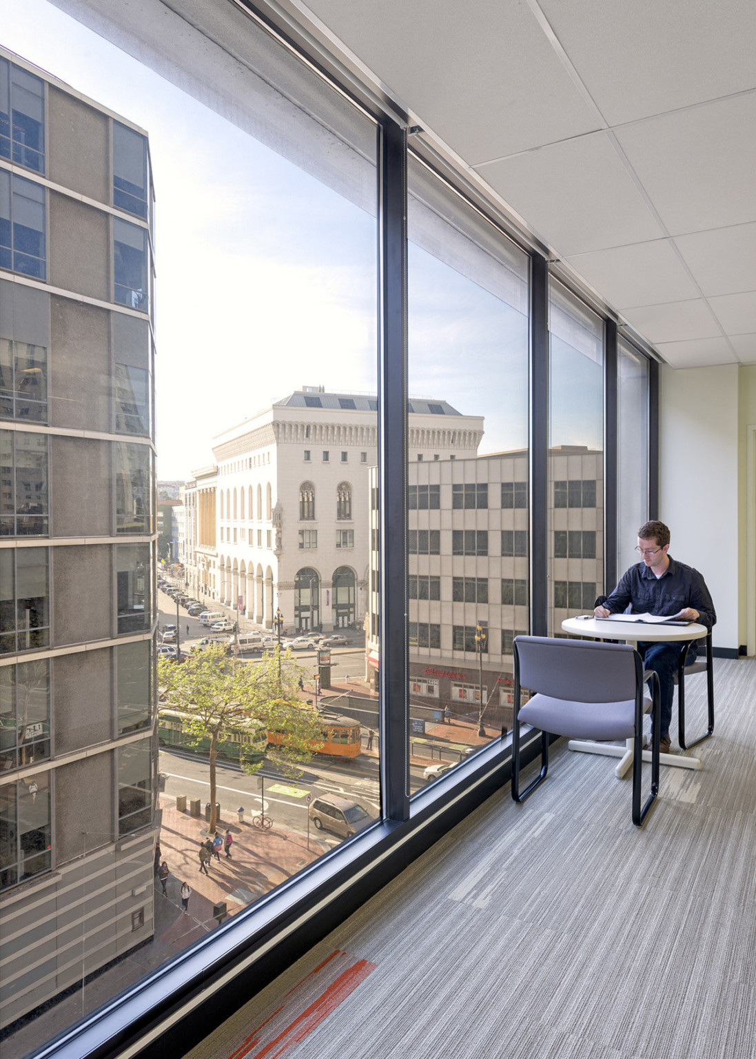 Table and chair in carpeted room next to floor to ceiling windows with view of city. White panel ceiling above