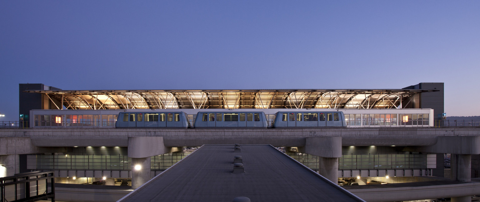 View from top of skywalk to train at station with illuminated curved half roof on opposite side with exposed beams.