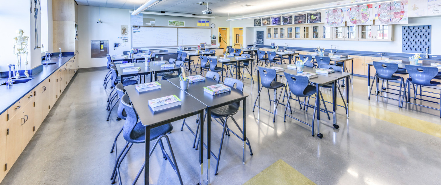 Interior classroom with flexible tables and blue chairs. Light wood cabinets with blue counters. Whiteboards on left wall