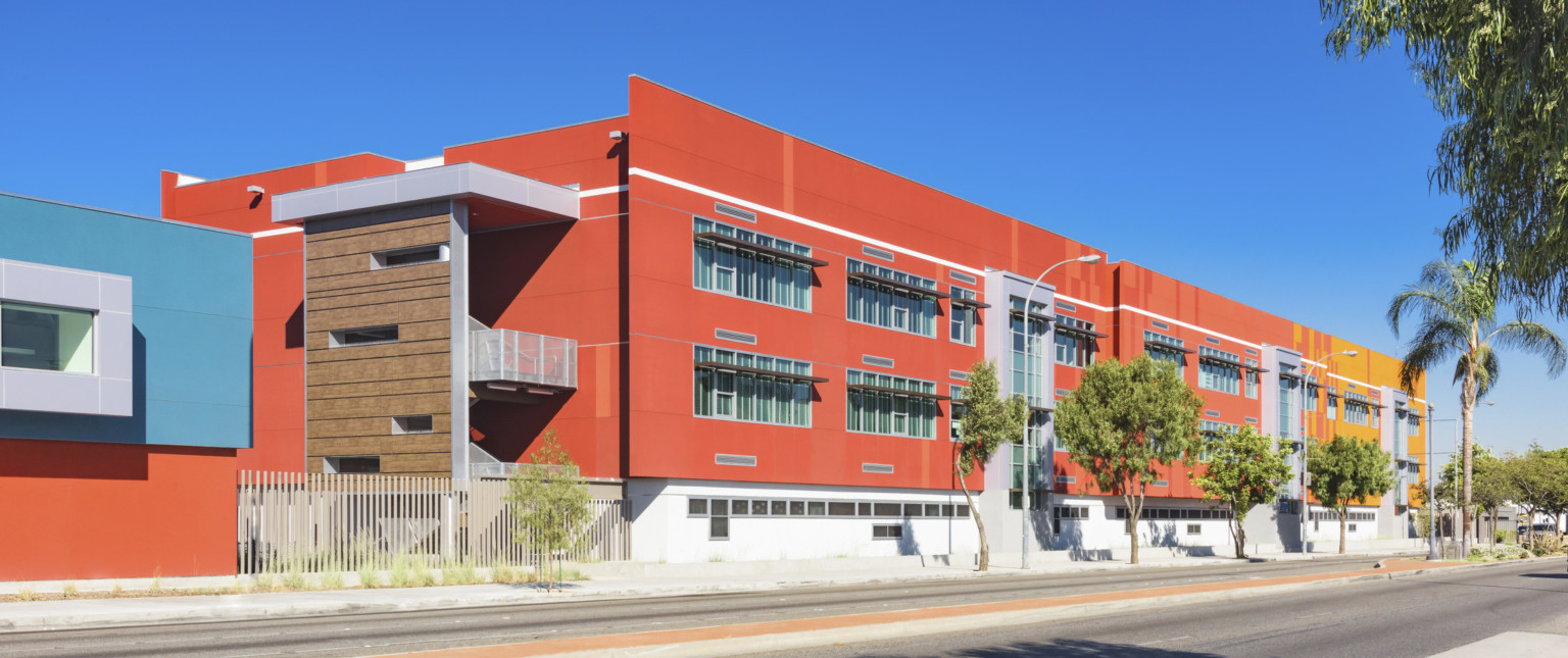 3 story red and yellow stucco building with exterior stairs, left, sheltered by wood panel wall. Silver wrapped facade accent