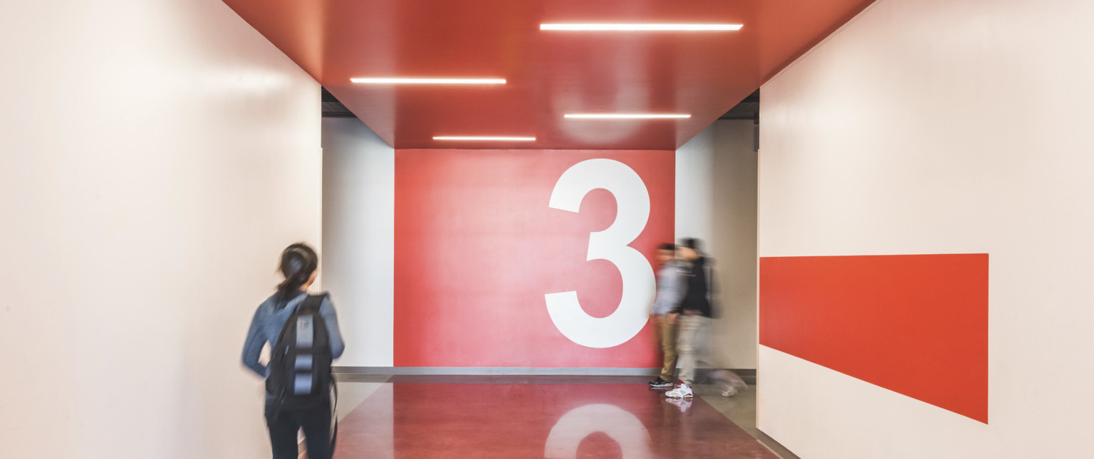 Intersection of two white hallways. Red tile on floor mirrors drop ceiling leading to red accent wall with 3 painted on