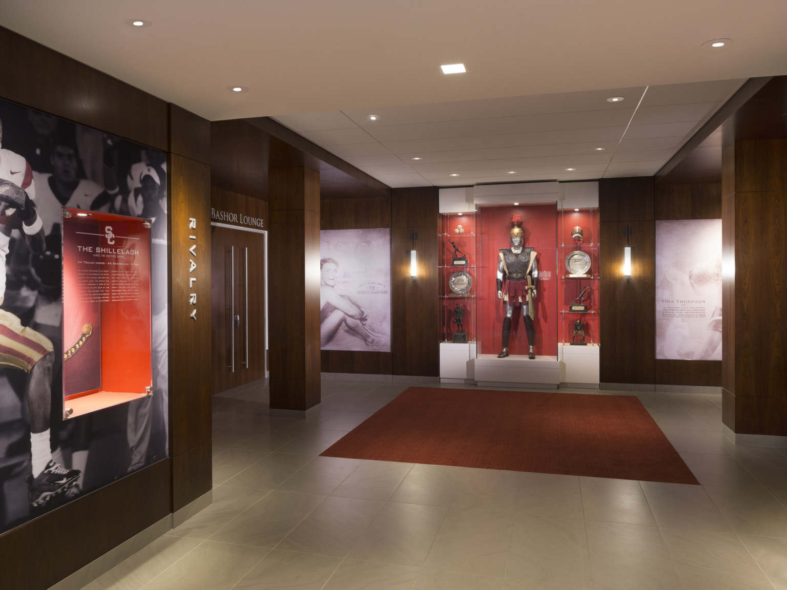 Display hallway with wood walls with murals and displays. White ceiling with recessed lights. Double doors to lounge, left