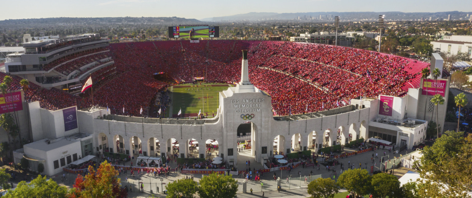 Front view of full stadium labeled Los Angeles Memorial Coliseum with Olympic Rings logo on central tower framed by archways