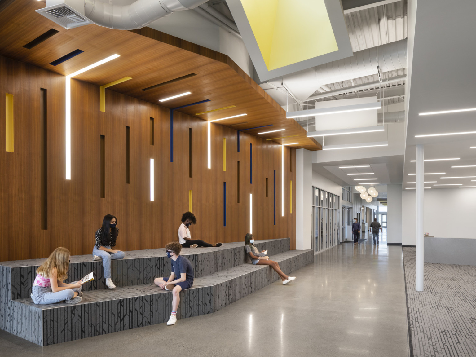 Grey stepped benches along wood wrapped accent wall with recessed lights and colored panels in white curving hallway