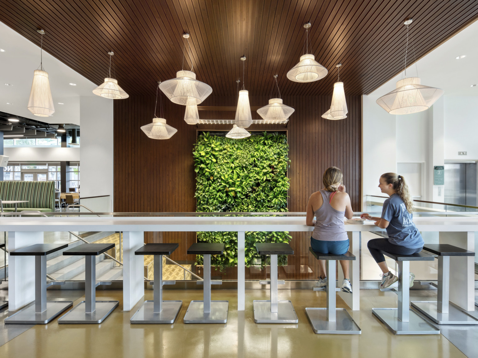 Counter and stools along glass railing over stairwell. Wood detail on stair wall and ceiling with living wall center