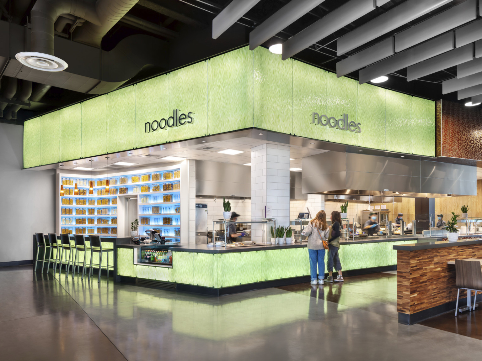 Illuminated green food station labeled noodles with bar seating left. Grey hanging panel details, exposed ducts along ceiling