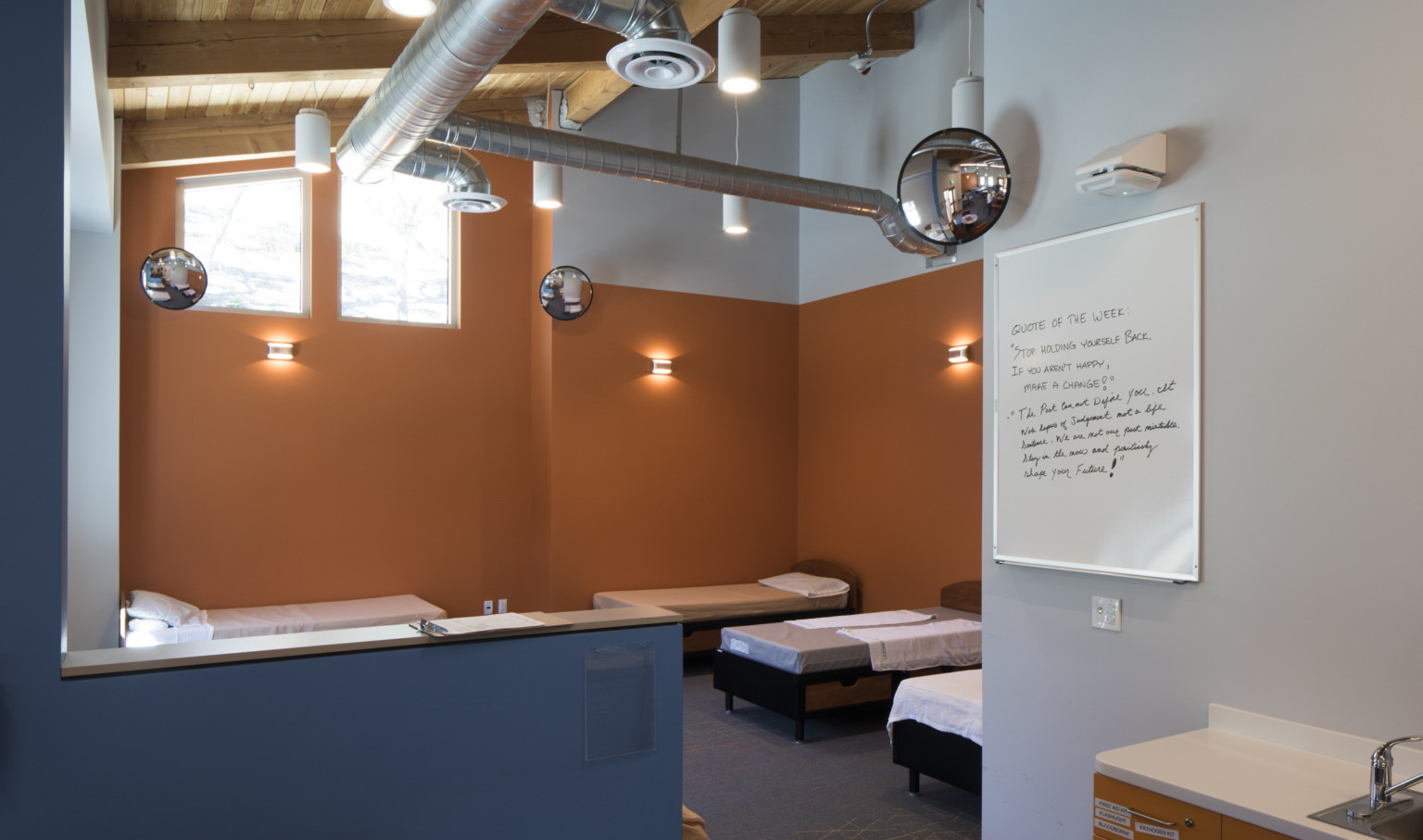 Orange bedroom with 6 beds separated from seating area by blue half wall. Exposed ductwork and round mirrors overhead