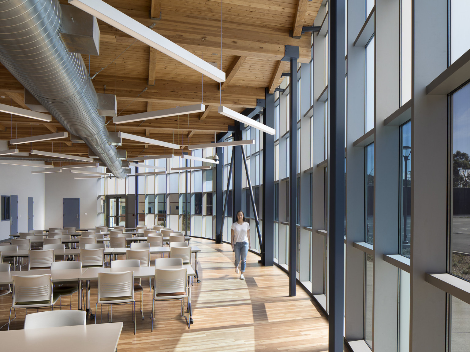 Interior view with curved glass wall, right. Left, a seating area with tables under wood ceiling with exposed beams and ducts