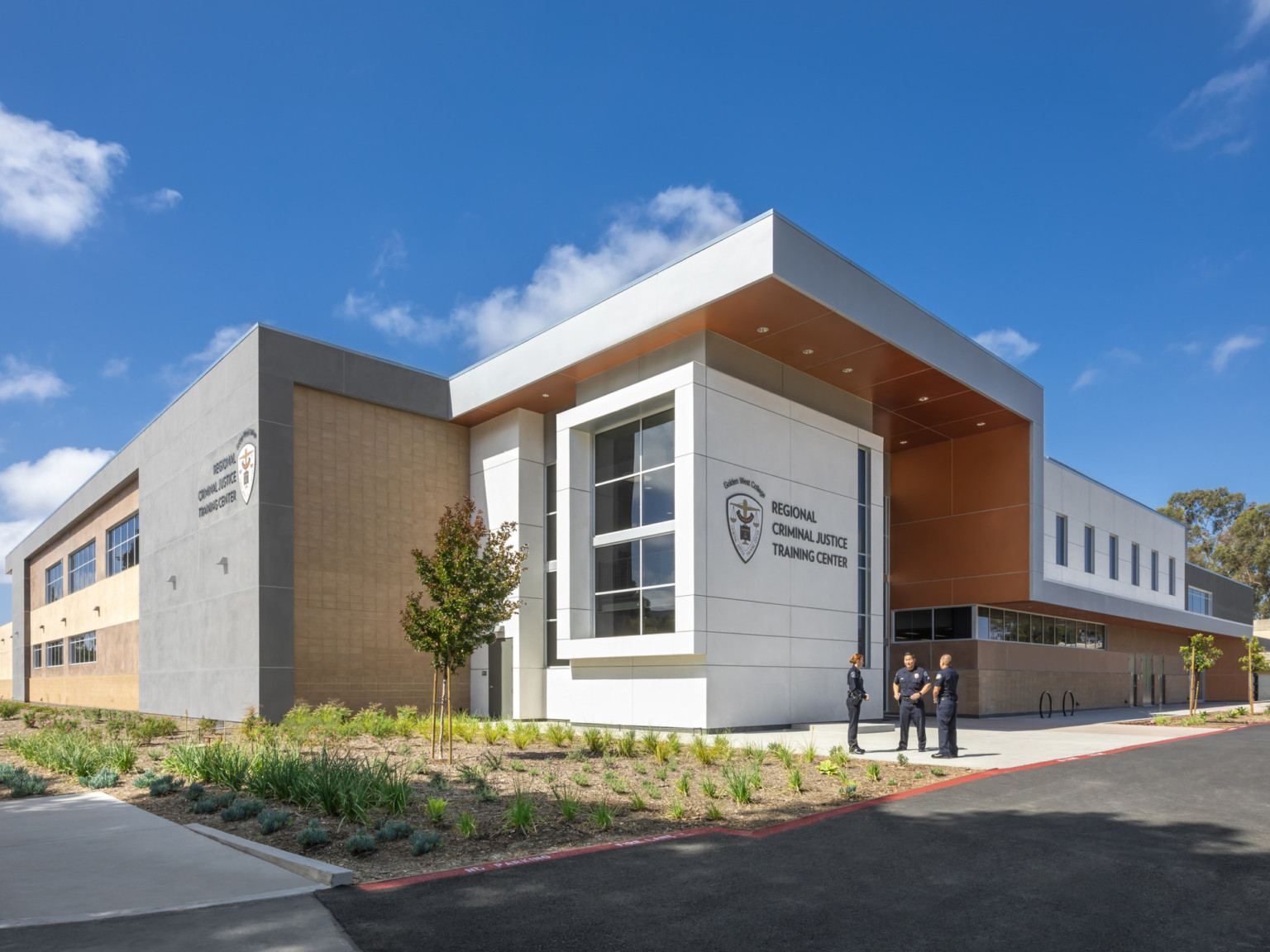 Corner exterior view of white cubic entrance with logo facing the street, shaded by a grey overhang with orange base