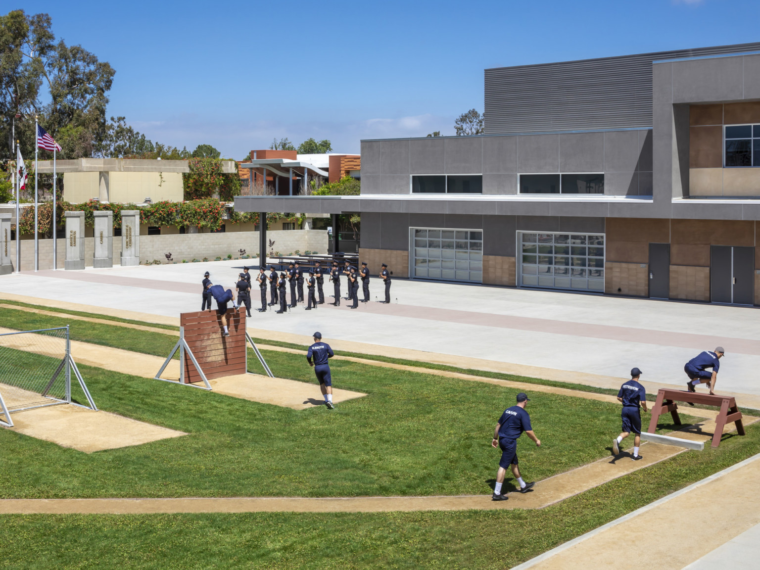 Outdoor training area with obstacles on course. In the background, people stand in front of staggered multistory building