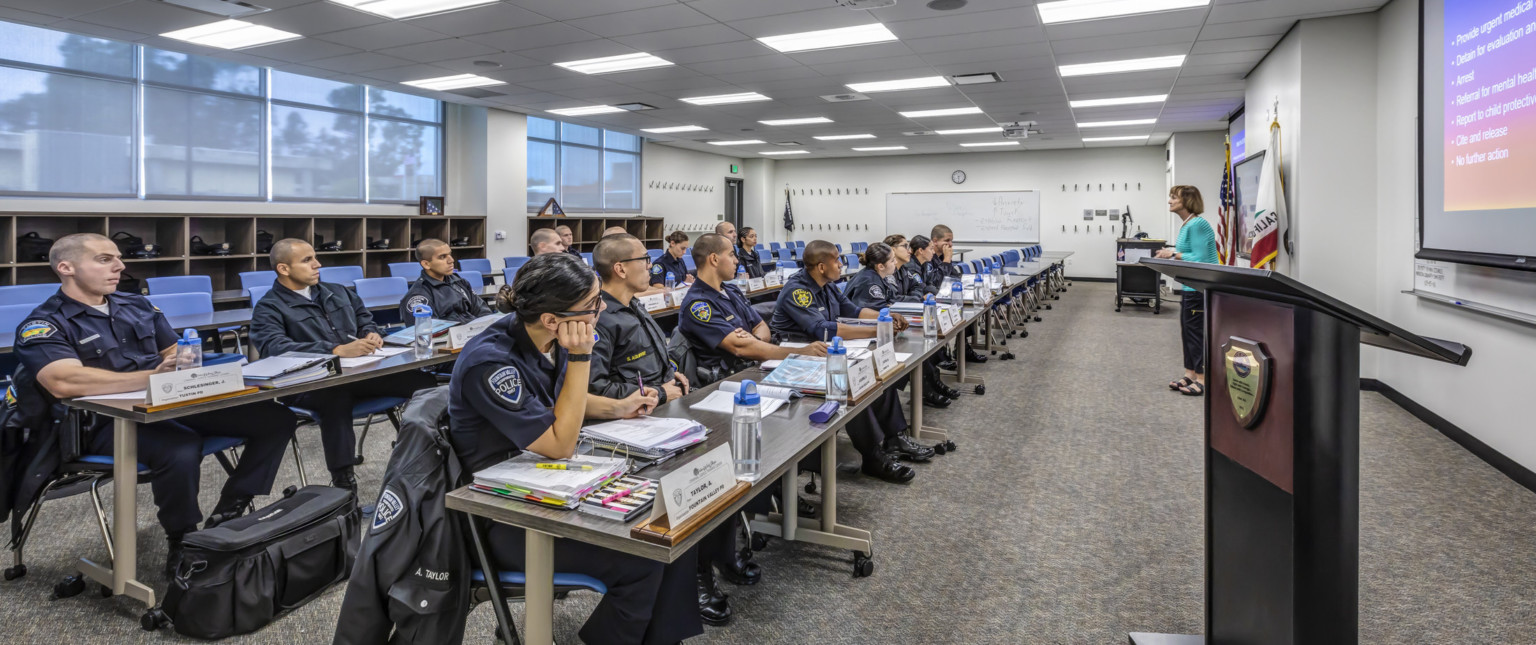 Officers sit in classroom with four long sections of rolling tables facing teacher at the front of the room