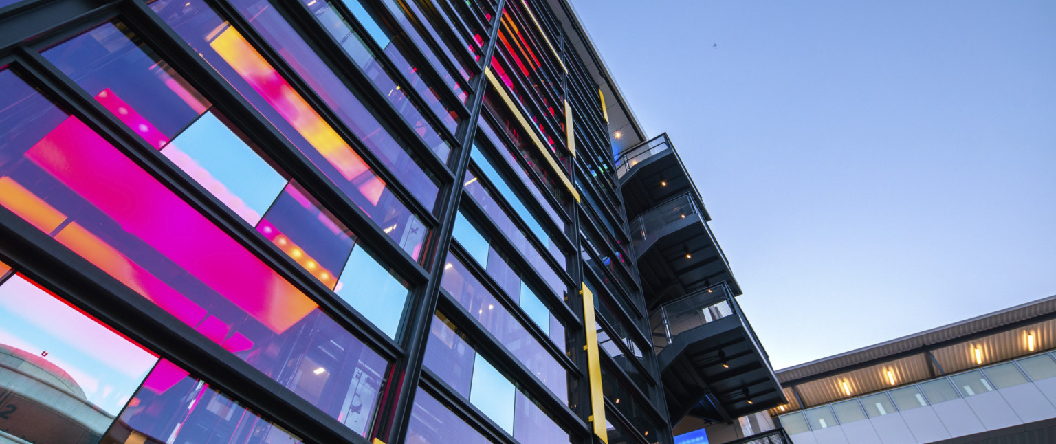 View looking up at central glass tower with iridescent window panels and staircase to the right. Skywalk over road to right