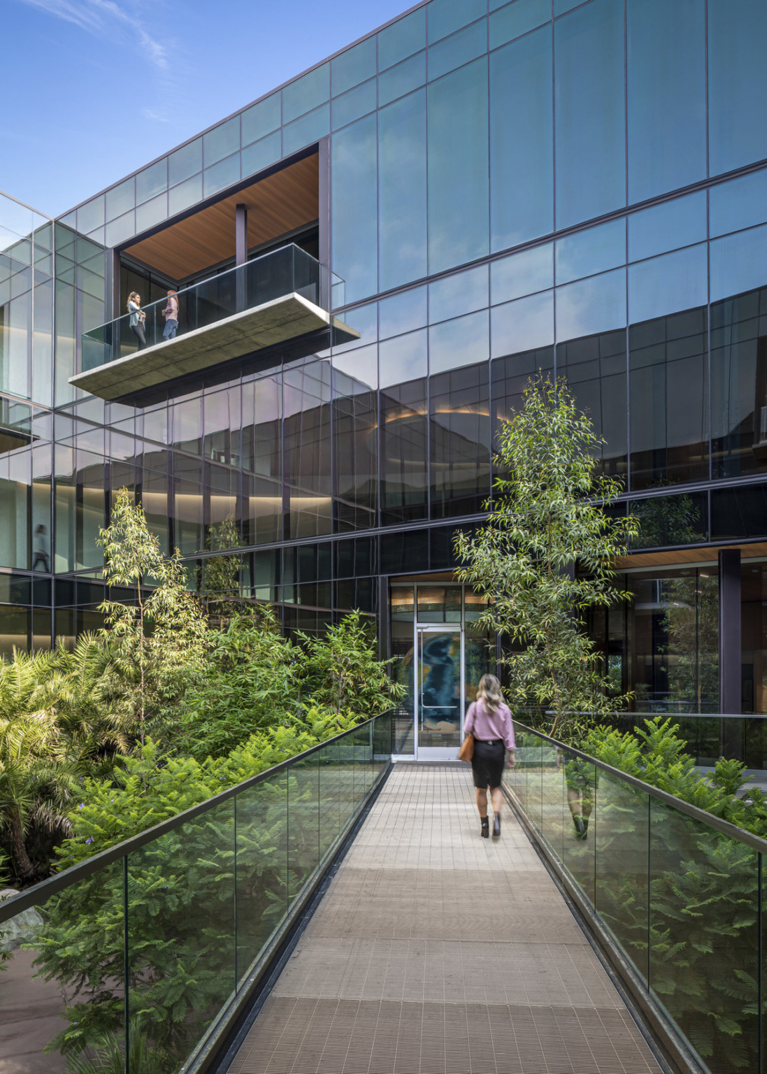 Pedestrian bridge with glass railings over courtyard to door of glass facade building with overhanging balcony