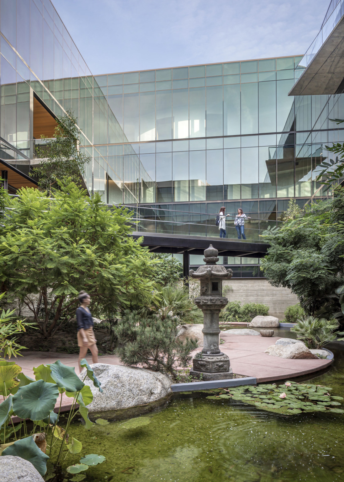 View of courtyard with large green plants and statues from across pond. Pedestrian bridge, above, framed by glass building.