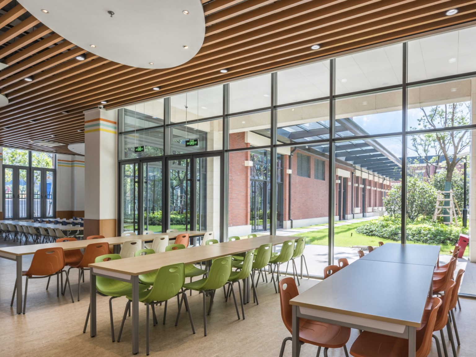 Dining area with long tables and colorful chairs looking out from floor to ceiling windows onto a courtyard with trees