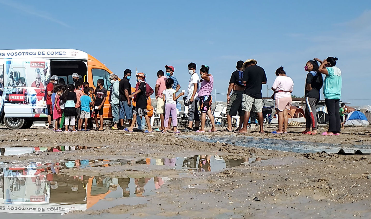 Line of people stand in muddy open space. Line leads to food donation van with side doors opened. Tents visible behind