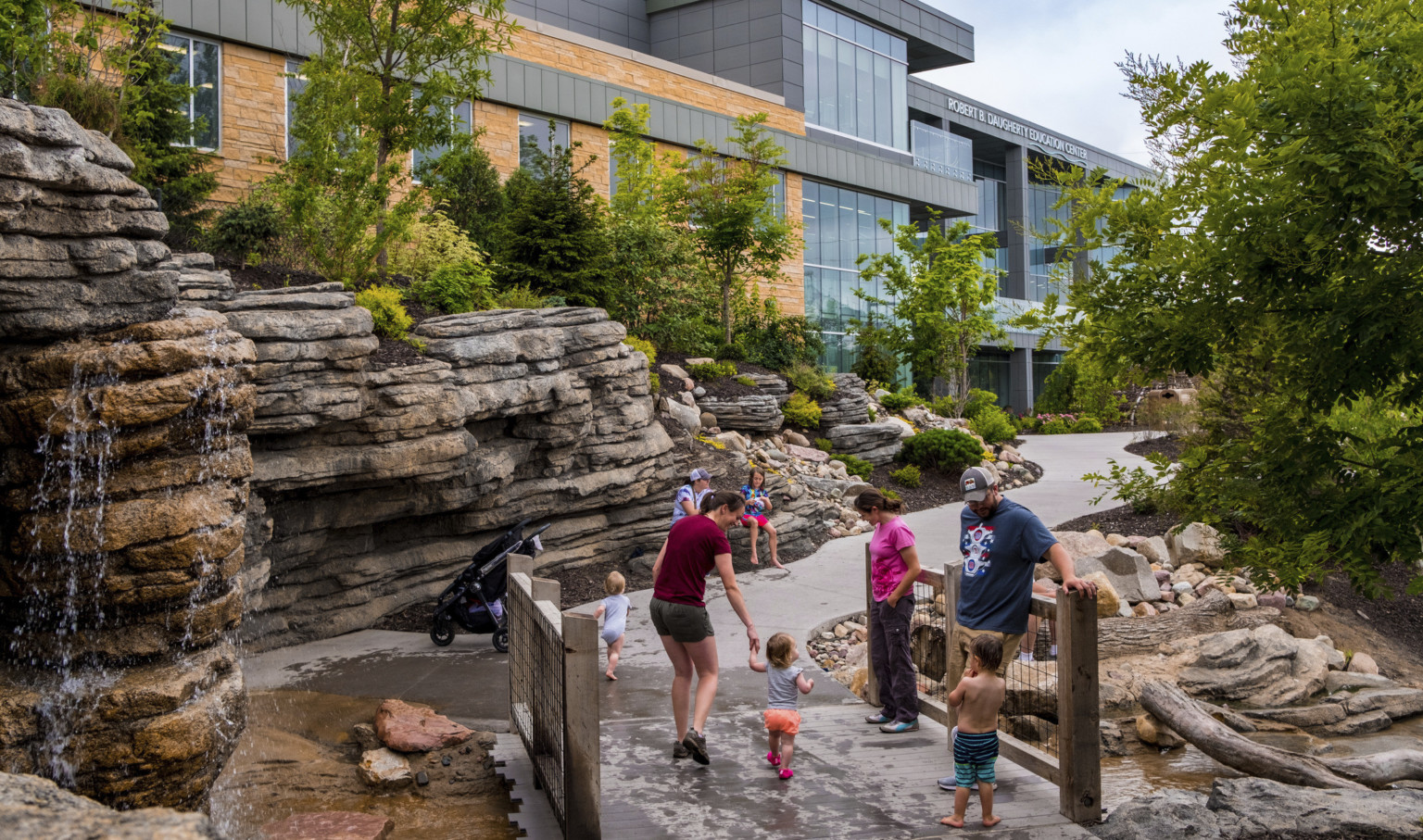 Small bridge over creek with waterfall flowing down rocks on the left along pathway leading to stone and glass building