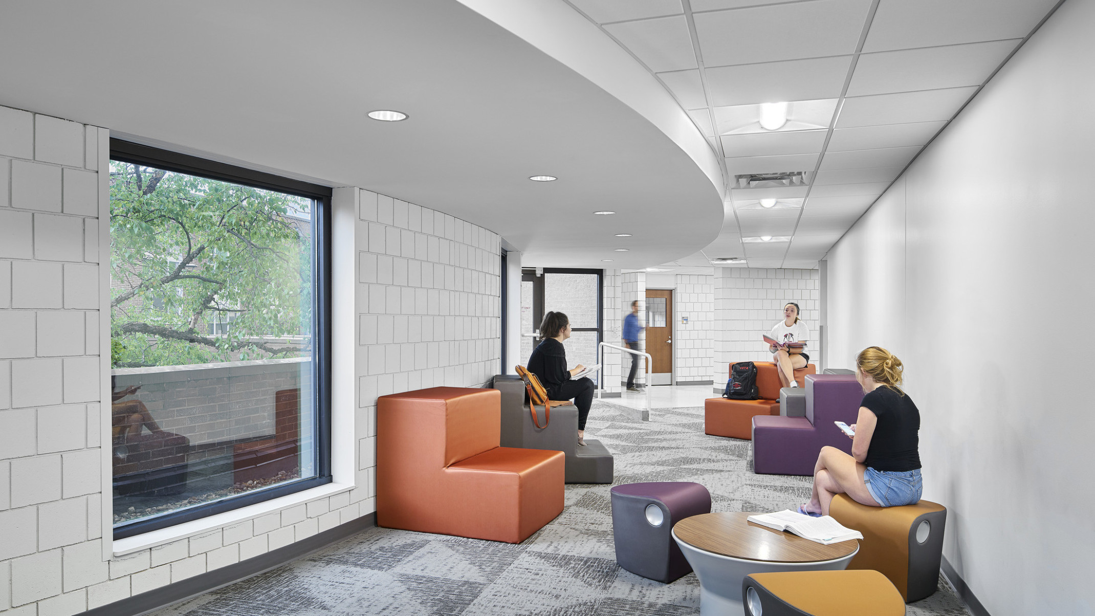 Renovated curved white tiled hallway for student interaction with colorful moveable seating. Curved drop ceiling detail, left