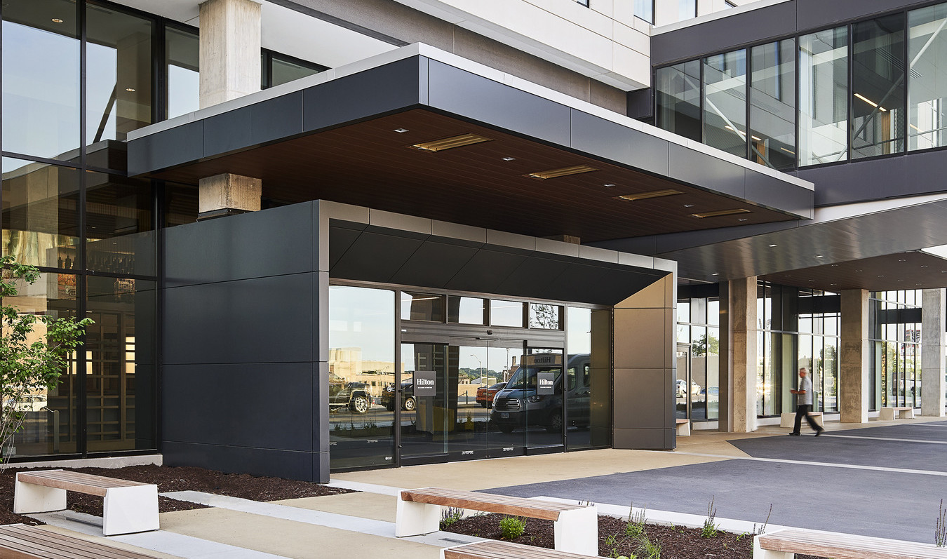 Entryway with benches out front, an overhang, and glass doors wrapped in dark framing at hilton des moines