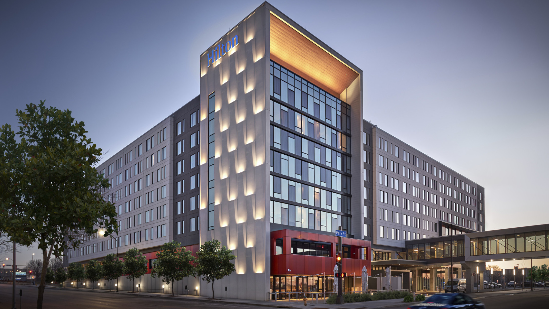 exterior corner view in the evening of multistory hotel with lit up roof arch, warm red entryway, and scaled facade with uplighting at hilton des moines