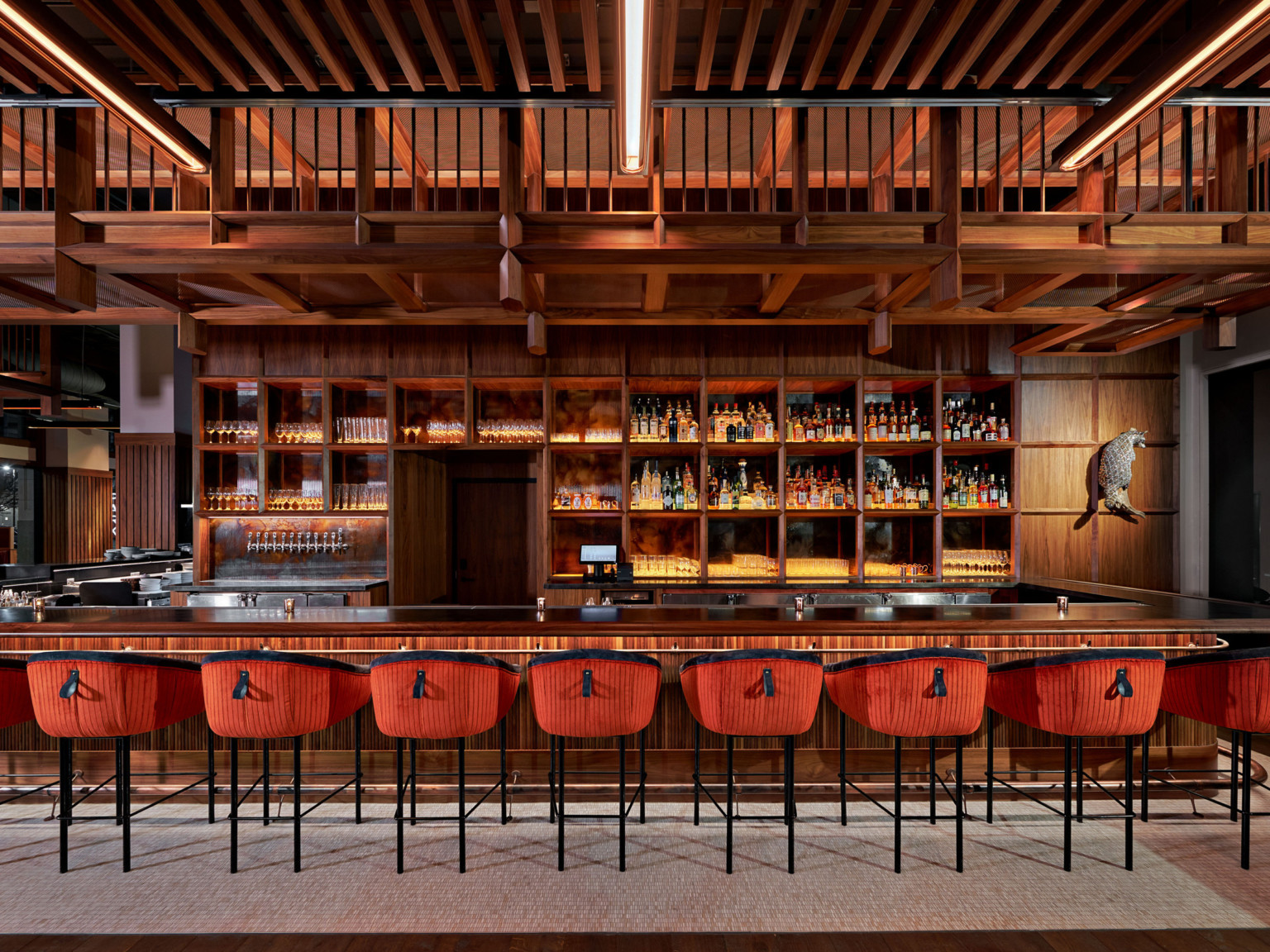 Large wood bar with built in shelves along the back. Wood overhang over back of bar, and stools with red and black upholstery