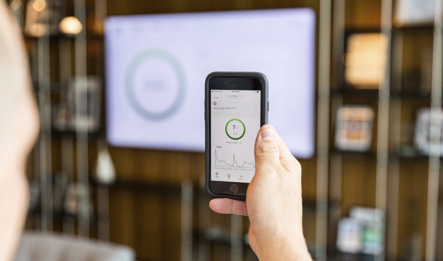 person using sonrai IAQ app in a space to read live air quality metrics, which are also shown on the lobby screen in front person using sonrai IAQ app in a space to read live air quality metrics, which are also shown on the lobby screen in front