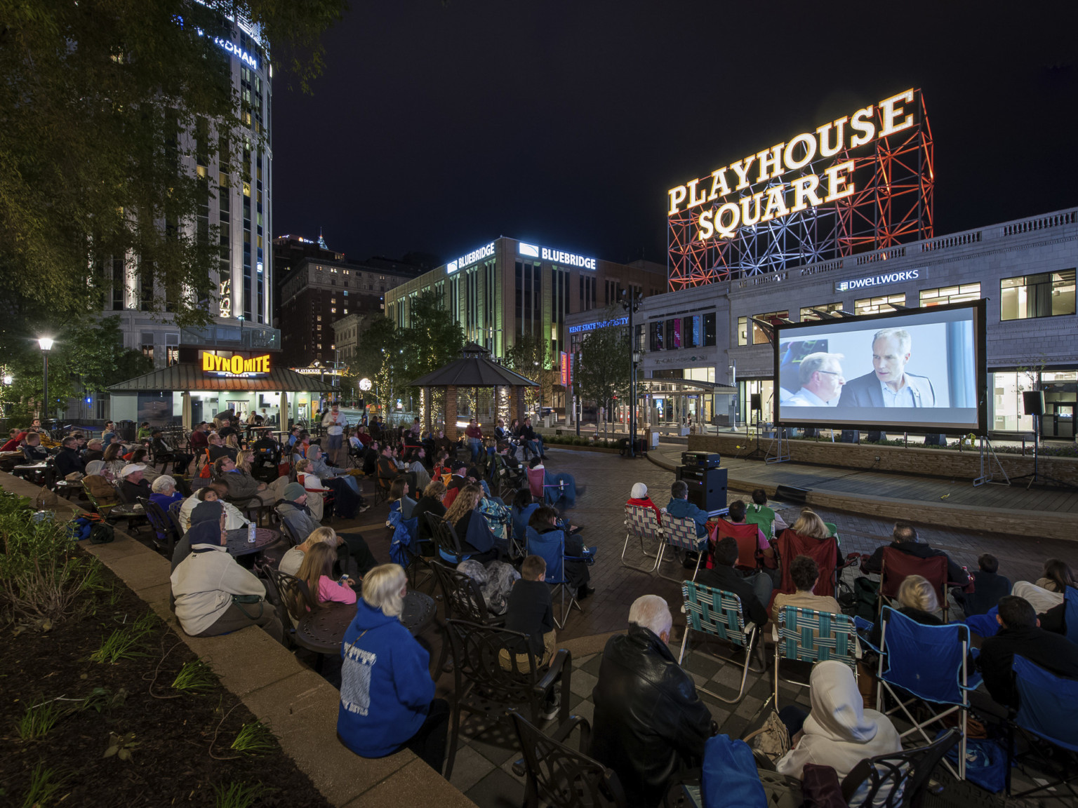 Audience sits on the street watching an outdoor movie on a screen. A Playhouse Square sign is illuminated in the background