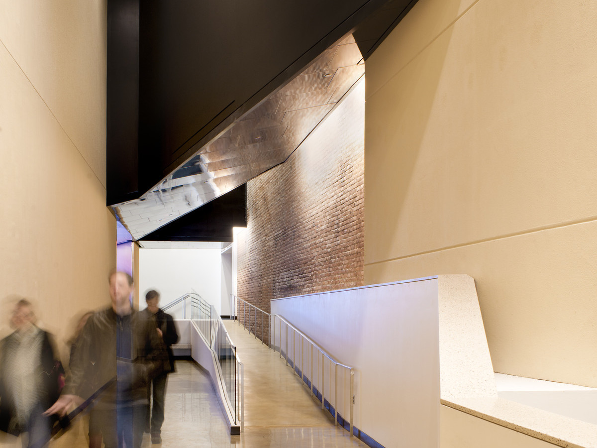 Hallway with ramp to the right, concrete yellow walls with exposed brick section reflected on metal ceiling section