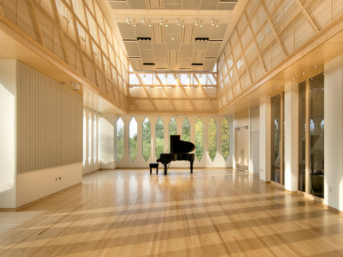 Piano in the center of a corner room with white walls and thin rectangular windows with pointed tops and bottoms, wood floor