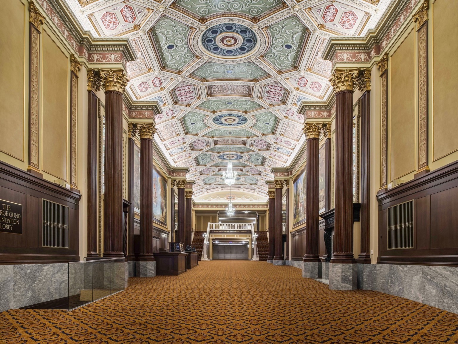 Grand staircase carpeted beneath ornate multicolored coffered ceiling with chandelier framed by ribbed pillars