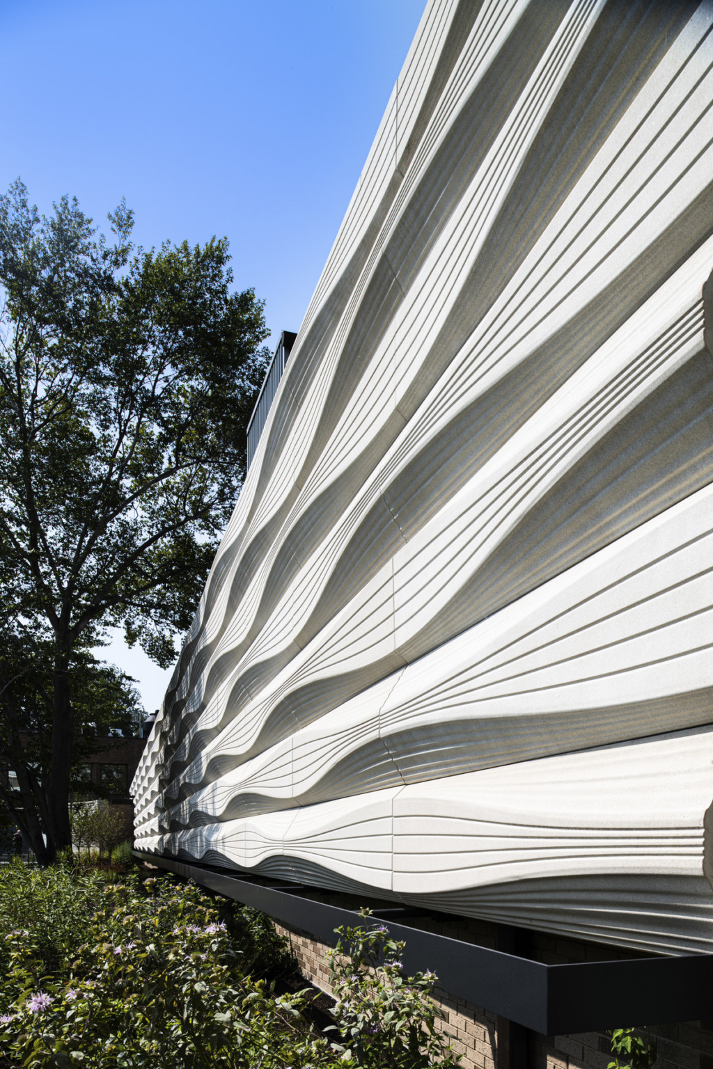 A white three dimensional building facade with horizontal striations and curves sit atop a brick plinth. Greenery surrounds.