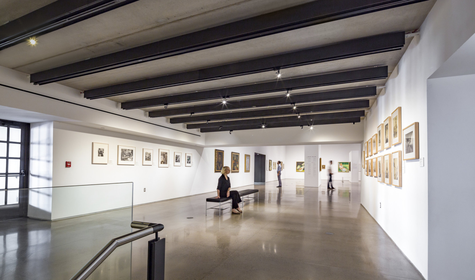 White gallery hall with paintings on the walls and benches along the center. Black beams cross the room's width with lighting