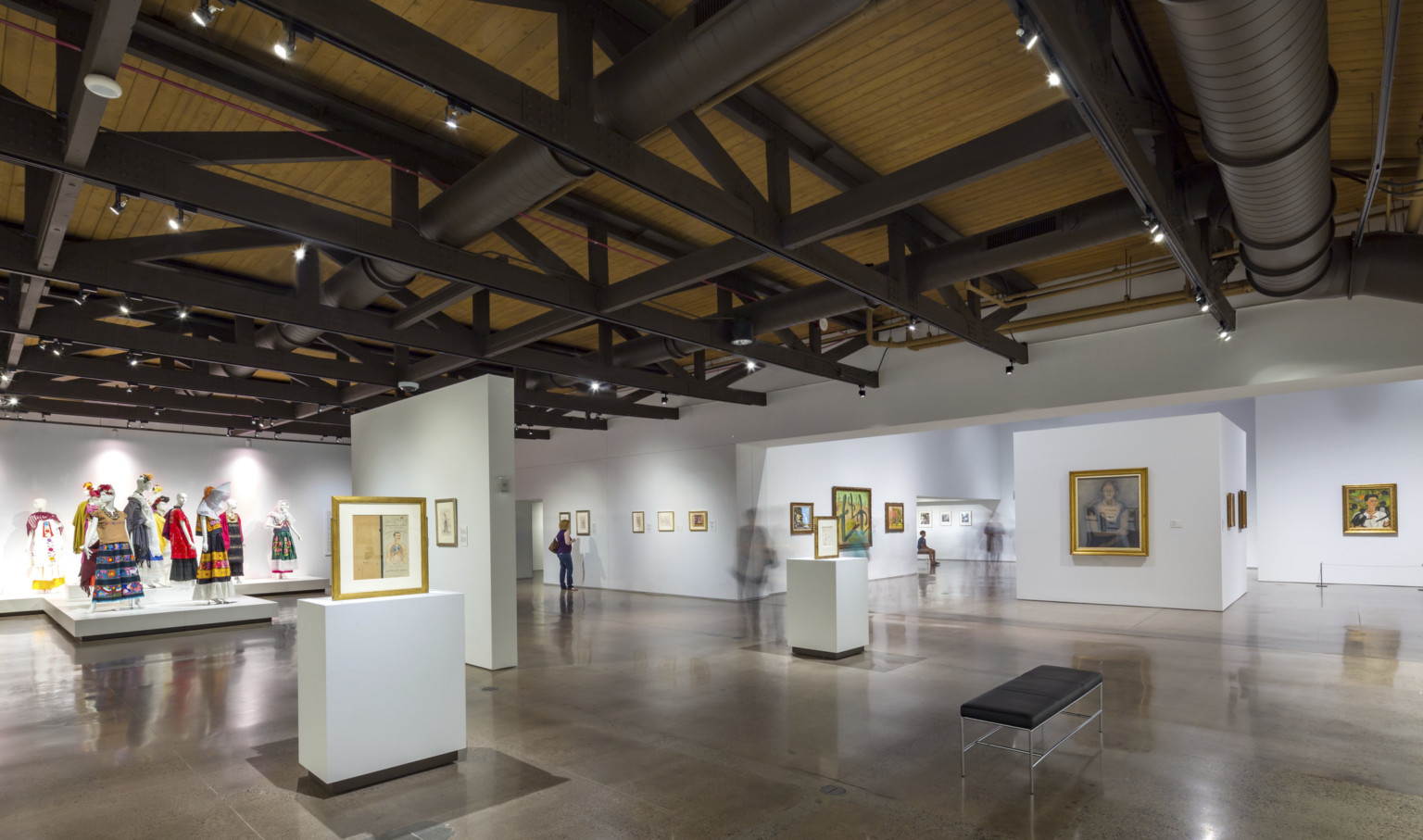 A gallery with paintings and a display of dressed mannequins, under a wood ceiling with exposed black support beams and pipes