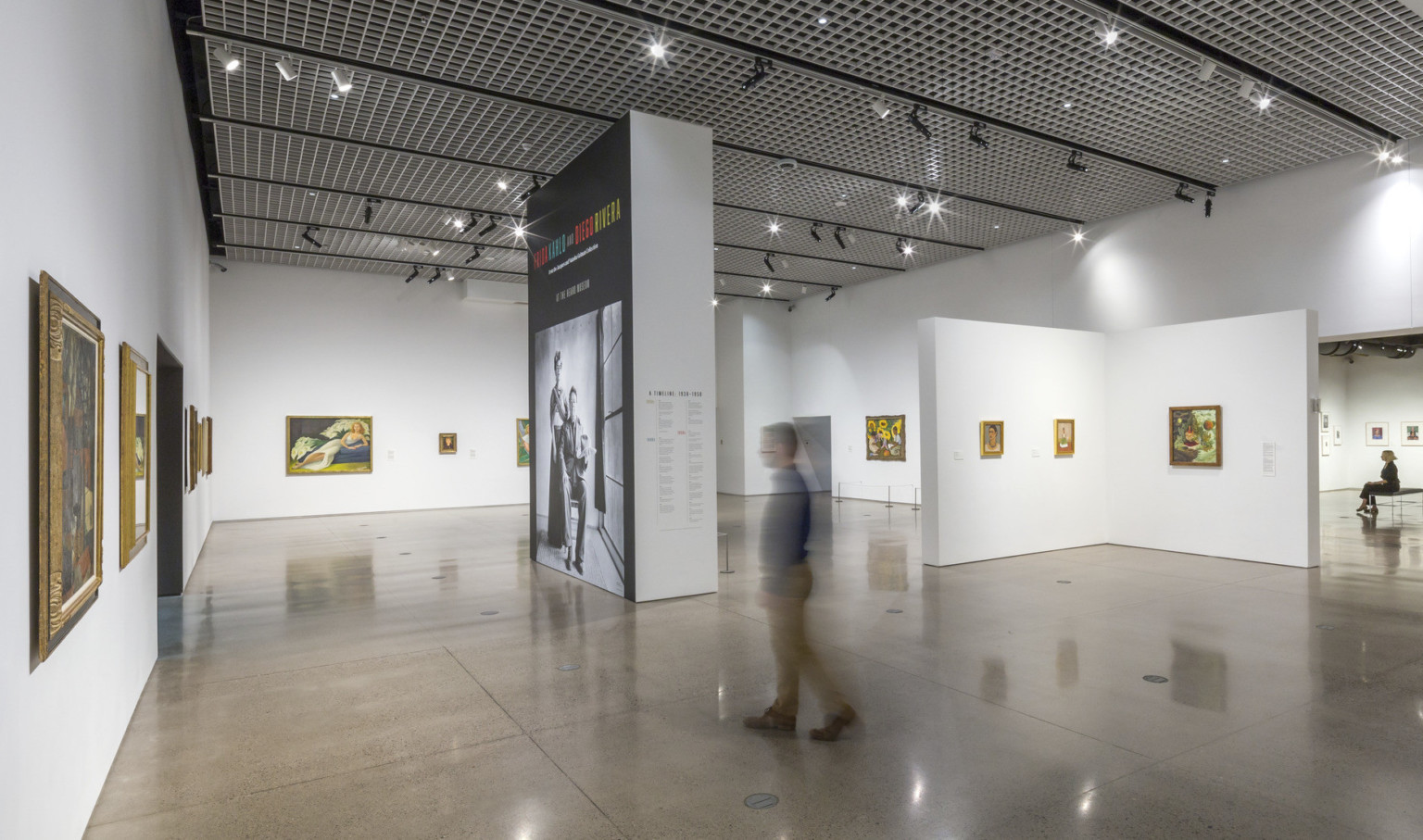 A white gallery in the Heard Museum with paintings displayed on the walls and a view of the adjoining gallery