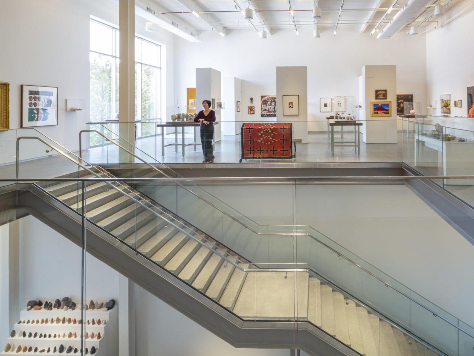 view from second story of art gallery split by staircase with white walls and custom lighting solution illuminating exhibits