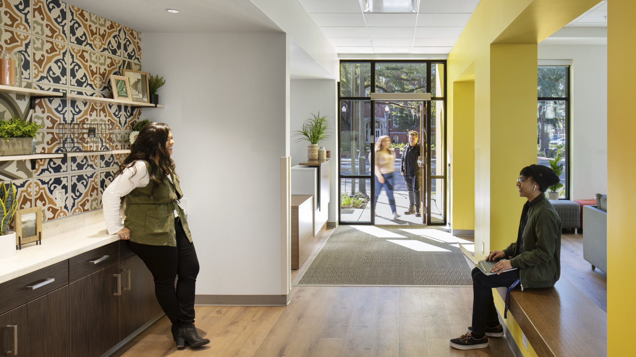 Hallway left of yellow wall to entry. White room towards the door behind recessed dark wood counter with muraled backsplash