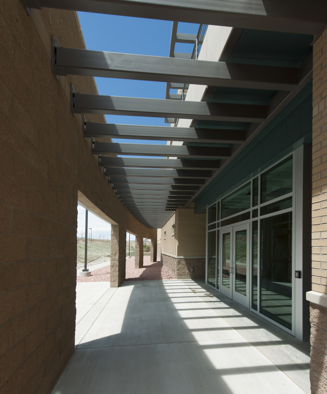 concrete walkway along a brick building and glass front entry shaded by metal fin canopy. blue sky above