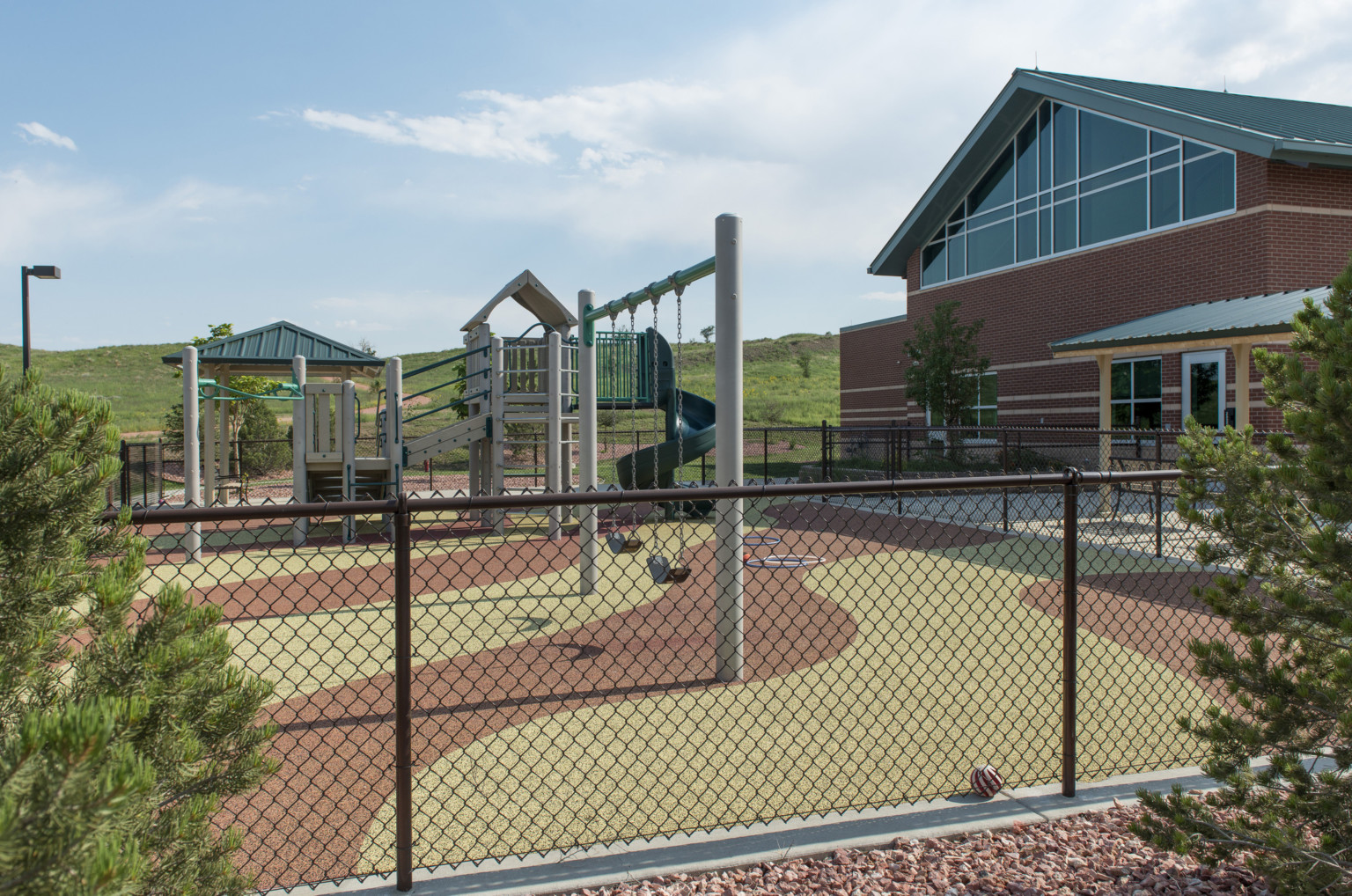 green and red poured in place rubber playground with swing set and climbing structure. brick building with peaked blue roof